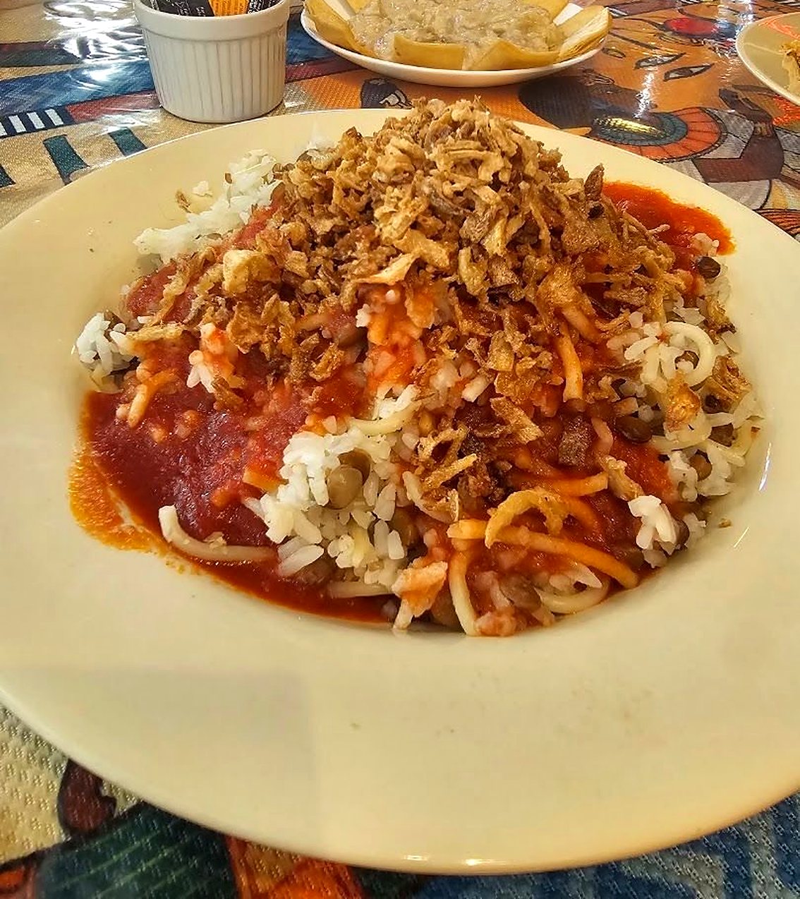 A plate of koshari: rice, macaroni and lentils under tomato sauce, topped with a heap of crispy fried onions, on a colourful patterned tablecloth