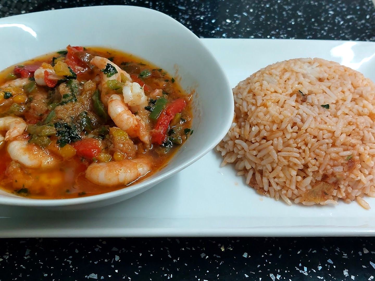 A bowl of prawns cooked with tomato, peppers and greens, next to a mound of tomato rice, on a white plate
